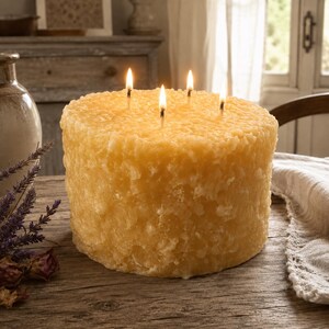 May include: A large, cylindrical, golden-yellow beeswax candle with a textured, uneven surface. The candle features four lit wicks positioned on its top flat surface, emitting a warm glow. The wax has a granular, honeycomb-like appearance.