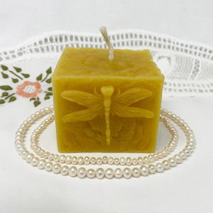 May include: A square, yellow beeswax candle with a dragonfly design. The candle has a white wick and is sitting on a white surface.