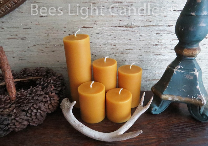 May include: Five beeswax candles of varying sizes, arranged in a row on a wooden surface. The candles are a warm, honey-colored yellow. A white antler is placed in front of the candles. The background is a rustic, wood-grain wallpaper with the text "Bees Light Candles" printed at the top.
