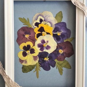May include: A framed pressed flower art piece featuring a bouquet of purple, yellow, and white pansies on a blue background.