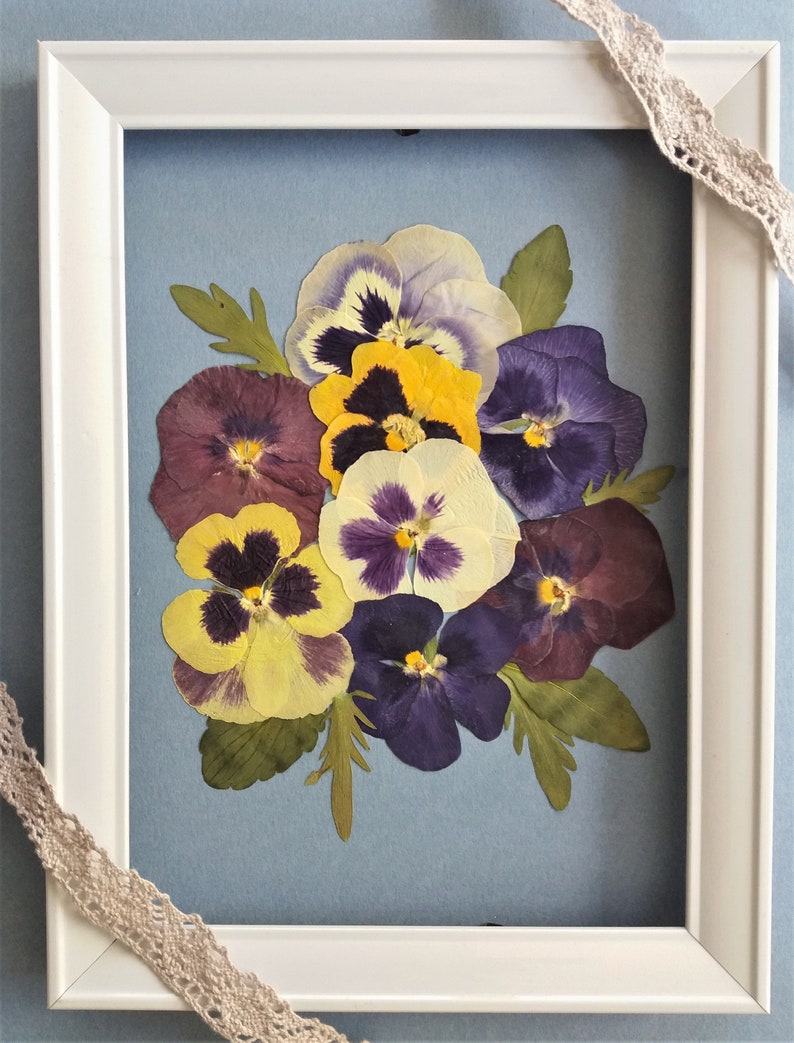 May include: A framed pressed flower art piece featuring a bouquet of purple, yellow, and white pansies on a blue background.