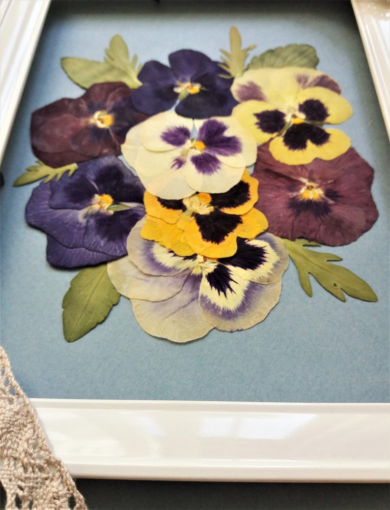 May include: A framed pressed flower arrangement featuring a bouquet of purple, yellow, and white pansies.