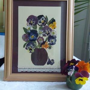 May include: A framed pressed flower art piece featuring a bouquet of purple, white, and yellow pansies in a brown vase. The frame has a white lace trim and a brown mat.