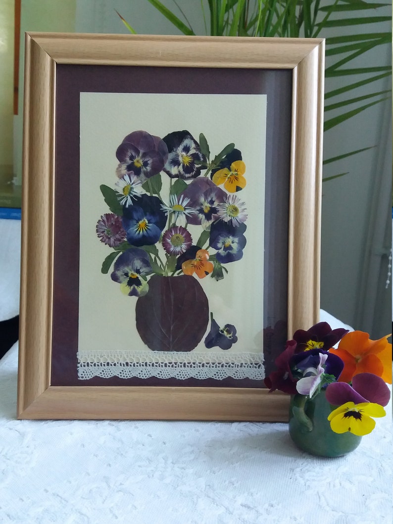 May include: A framed pressed flower art piece featuring a bouquet of purple, white, and yellow pansies in a brown vase. The frame has a white lace trim and a brown mat.