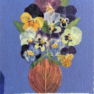 May include: A handmade card featuring a bouquet of pressed pansies in shades of purple, yellow, and blue arranged in a brown vase. The card is decorated with a white lace trim and a blue ribbon.