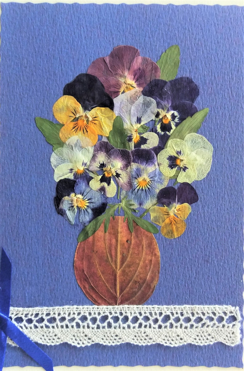 May include: A handmade card featuring a bouquet of pressed pansies in shades of purple, yellow, and blue arranged in a brown vase. The card is decorated with a white lace trim and a blue ribbon.