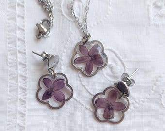 May include: A silver necklace and earrings set with a flower pendant and matching earrings. The flower is pressed and preserved in clear resin. The flower is purple.
