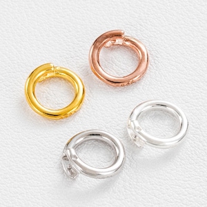 Può includere: Quattro piccole chiusure rotonde in oro, oro rosa e argento. Le chiusure sono aperte, con una piccola cerniera e un meccanismo di chiusura. La chiusura dorata ha piccoli abbellimenti scintillanti. Le chiusure sono su una superficie bianca.