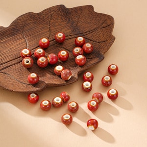 May include: A collection of red ceramic beads with speckled surfaces and white centers. The beads are scattered on a wooden leaf-shaped tray, with a neutral background. These beads are suitable for jewelry making and crafting projects.