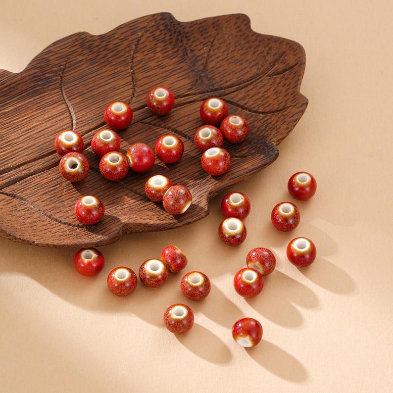 May include: A collection of red ceramic beads with speckled surfaces and white centers. The beads are scattered on a wooden leaf-shaped tray, with a neutral background. These beads are suitable for jewelry making and crafting projects.
