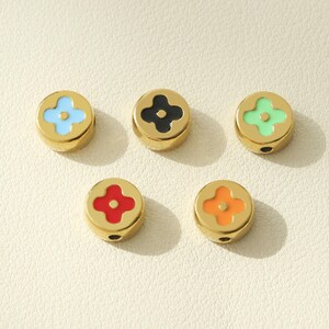 May include: Five gold-coloured round beads with a four-petal flower design in the centre. The beads are in different colours: light blue, black, light green, red, and orange.
