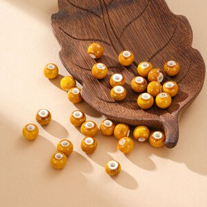 May include: A collection of yellow ceramic beads with white centers scattered on a wooden leaf-shaped tray. The beads have a glossy finish and are arranged on a light beige surface.