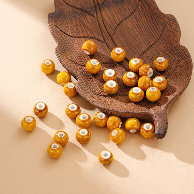 May include: A collection of yellow ceramic beads with white centers scattered on a wooden leaf-shaped tray. The beads have a glossy finish and are arranged on a light beige surface.