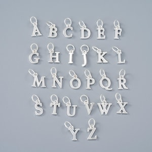 May include: A set of silver alphabet charms, each with a small loop for attaching to jewellery. The letters A to Z are displayed, all in a simple, sans-serif font. The charms are arranged on a grey background.