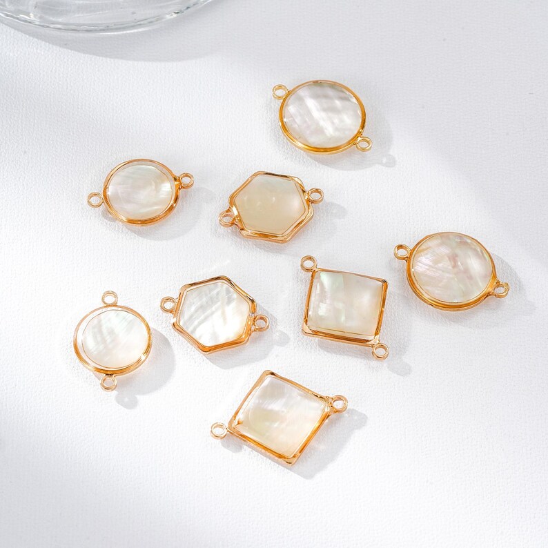 May include: A collection of gold-toned jewelry components with iridescent, shell-like inserts. The components feature various shapes, including circles, hexagons, and squares, each with small loops for attachment. The pieces are arranged on a white surface.