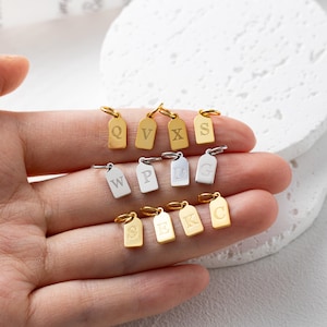 Op de afbeelding: Een verzameling kleine, tag-vormige hangers met verschillende gegraveerde letters. De hangers zijn in goud- en zilverkleuren, elk met een kleine lus voor bevestiging. De letters zijn Q, V, X, S, W, P, D, G, E, K en C.