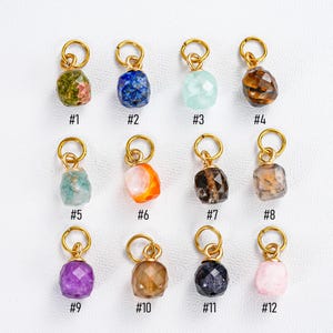 May include: A collection of twelve gemstone charms with gold-colored loops. The gemstones vary in color, including green, blue, clear, brown, orange, purple, and pink. Each charm is numbered from #1 to #12.