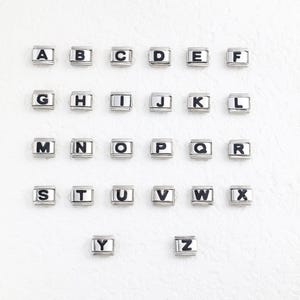 Puede incluir: Abalorios plateados con letras negras del alfabeto, dispuestos sobre un fondo blanco. Cada abalorio es cuadrado con un marco rectangular. Se muestran las letras de la A a la Z.