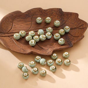 May include: A collection of small, round ceramic beads with a speckled green and brown glaze. The beads have a central hole and are displayed on a wooden, leaf-shaped tray. Ideal for jewelry making or craft projects.