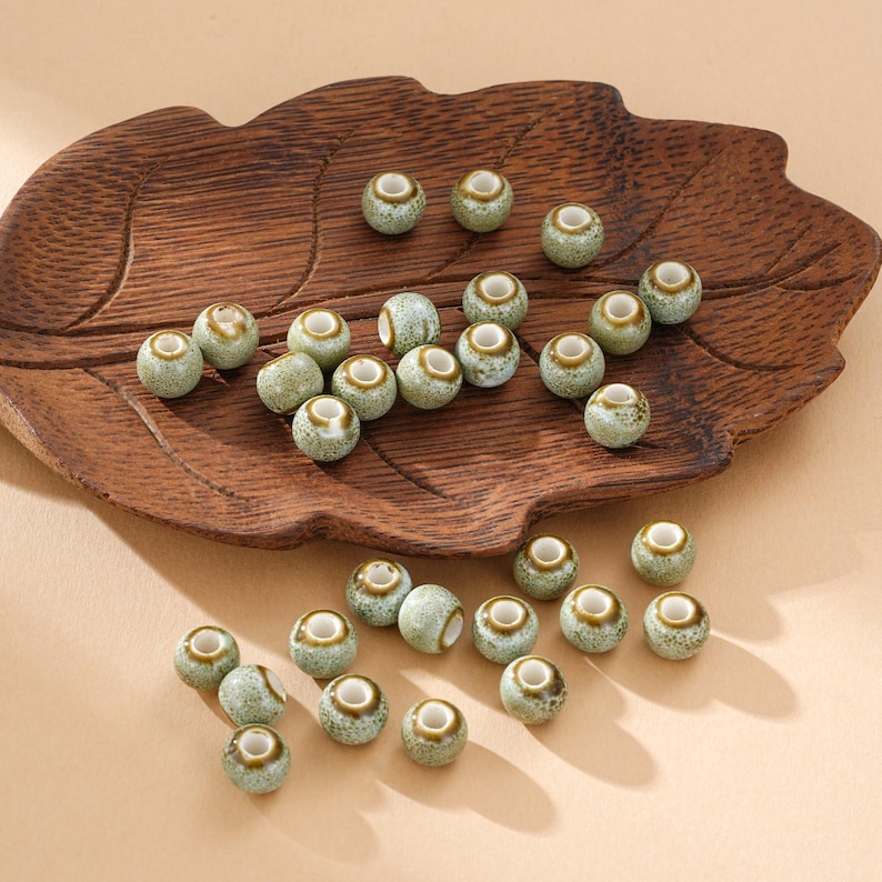 May include: A collection of small, round ceramic beads with a speckled green and brown glaze. The beads have a central hole and are displayed on a wooden, leaf-shaped tray. Ideal for jewelry making or craft projects.
