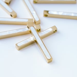 以下が含まれることがあります： 白いインレイが付いた金色の金属製のバーペンダント。ペンダントは十字形に配置されています。