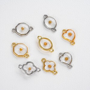 Pu&ograve; includere: Una collezione di ciondoli assortiti con un seme racchiuso in resina trasparente. I ciondoli hanno montature in metallo argentato e dorato di varie forme, tra cui un cuore, un ovale e un trifoglio. Lo sfondo &egrave; una superficie bianca.