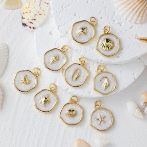 May include: A selection of gold-coloured pendants with white enamel backgrounds, each featuring a different sea-themed design. The pendants include a starfish, seashell, and turtle. The pendants are displayed on a white surface with seashells.