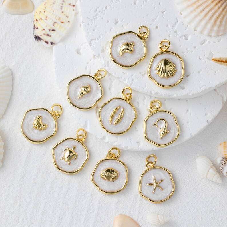 May include: A selection of gold-coloured pendants with white enamel backgrounds, each featuring a different sea-themed design. The pendants include a starfish, seashell, and turtle. The pendants are displayed on a white surface with seashells.