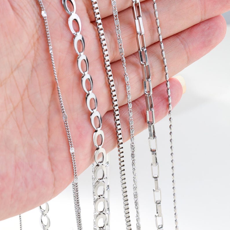 May include: A hand displays a variety of silver necklaces. The necklaces feature different chain styles, including oval links, box chains, and delicate beaded designs. The chains are all a bright, shiny silver color.
