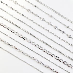 May include: An assortment of silver necklaces with various chain styles, including heart, cross, and oval link designs. The necklaces are displayed on a white surface, showcasing their different textures and patterns. The chains are made of a shiny, metallic material.