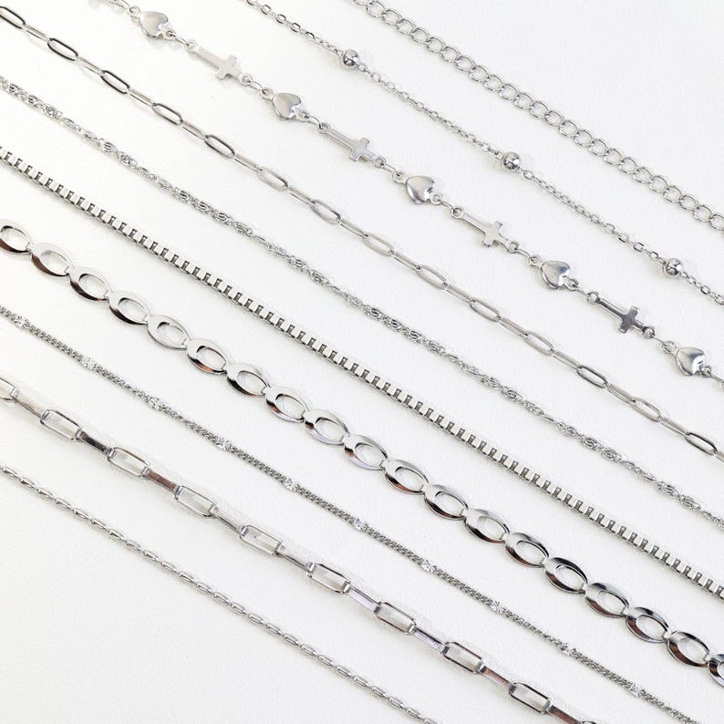 May include: An assortment of silver necklaces with various chain styles, including heart, cross, and oval link designs. The necklaces are displayed on a white surface, showcasing their different textures and patterns. The chains are made of a shiny, metallic material.
