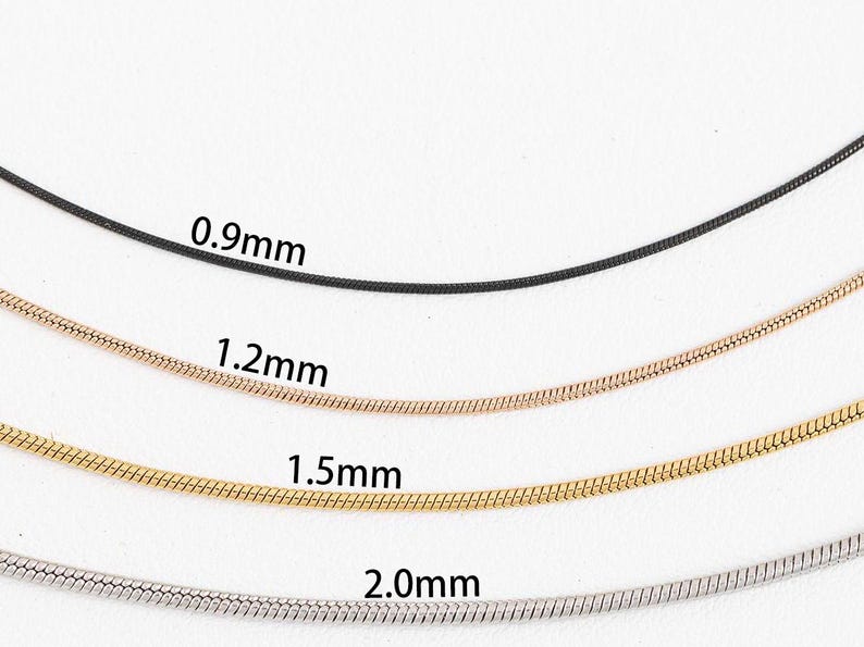 May include: Four snake chains of different thicknesses, from 0.9mm to 2.0mm, laid out on a white surface. The chains are black, rose gold, gold, and silver.