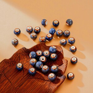 May include: A collection of blue ceramic beads with a white center and a brown rim. The beads are scattered on a wooden leaf-shaped tray. The beads are approximately 0.8 cm in diameter, perfect for jewelry making or craft projects.