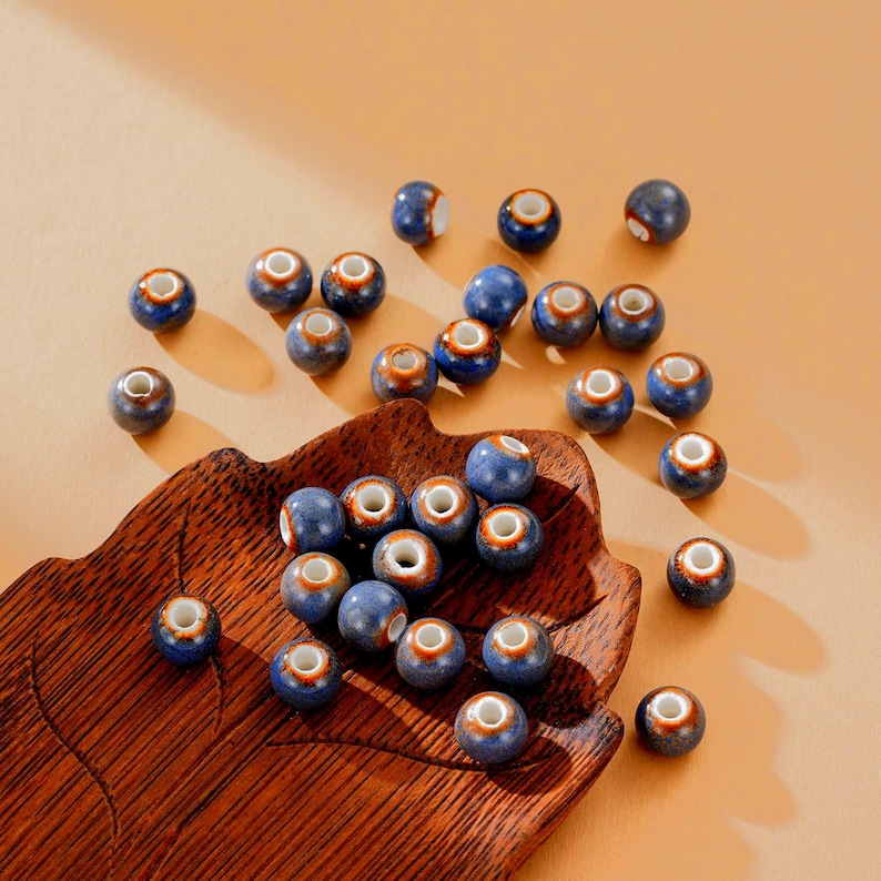 May include: A collection of blue ceramic beads with a white center and a brown rim. The beads are scattered on a wooden leaf-shaped tray. The beads are approximately 0.8 cm in diameter, perfect for jewelry making or craft projects.