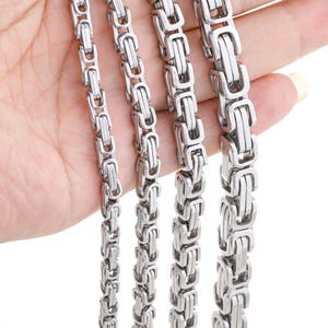 Puede incluir: Primer plano de varios collares de cadena bizantina de acero inoxidable. Las cadenas varían en anchura, mostrando el intrincado diseño de eslabones. Las cadenas plateadas se muestran sobre un fondo neutro, resaltando su acabado pulido. Estos collares son adecuados para el uso diario.