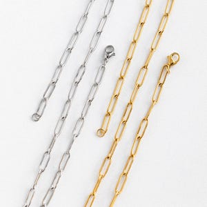 May include: Close-up of several paperclip-style chain necklaces in gold and silver. The chains have elongated oval links and are displayed on a white background. Some chains have clasps attached.