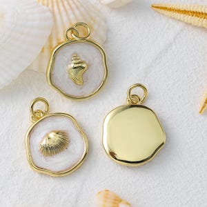 May include: Gold-coloured seashell pendants with a white enamel background. The pendants feature detailed seashell designs and a loop for attaching to a chain. The background is a pearlescent white. The pendants are on a sandy surface.