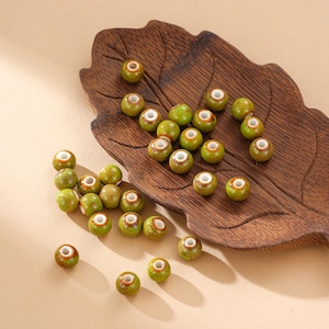 May include: A collection of small, round beads with a speckled green and brown finish. Each bead has a white center and a brown ring around the opening. The beads are scattered on a wooden leaf-shaped tray.