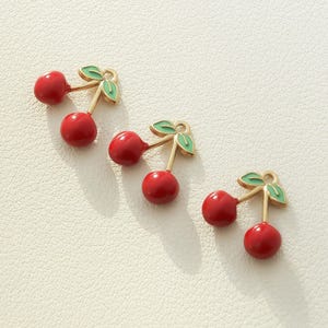 Op de afbeelding: Vier gouden kersenbedels met rood en groen emaille. Elke bedel heeft een kers met een groene steel en blad.