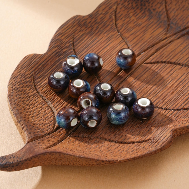 May include: A collection of ceramic beads with a glossy finish. The beads are primarily dark brown with blue and white accents. They are arranged on a wooden leaf-shaped tray, showcasing their round shape and unique color variations.