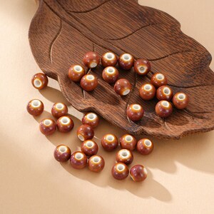 May include: A collection of small, round, brown ceramic beads with white centers. The beads are scattered on a wooden, leaf-shaped tray. The beads have a glossy finish and are suitable for jewelry making or crafts.