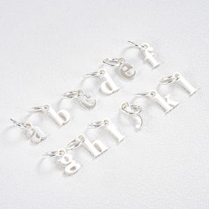 May include: A collection of small, silver-colored alphabet charms, each with a loop for attaching to a chain or bracelet. The letters a, b, c, d, e, f, g, h, j, and k are visible. The charms are arranged on a white surface.