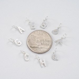May include: A collection of small, silver letter charms, each with a loop for attaching to a chain or bracelet. The letters A, B, C, D, E, G, H, K, and Q are visible. A U.S. quarter is included for size comparison.