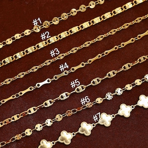 Può includere: Una selezione di sette collane a catena color oro, ciascuna con un design unico. Le catene sono esposte su una superficie marrone. I design includono maglie rotonde, maglie strutturate e ciondoli a forma di trifoglio. Ogni catena è numerata.