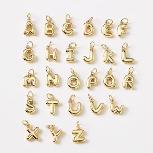 Puede incluir: Una colección de colgantes de alfabeto dorados, cada uno con un pequeño lazo para sujetar a una cadena o pulsera. Las letras están en una fuente redondeada, tipo burbuja, y están dispuestas en filas y columnas.