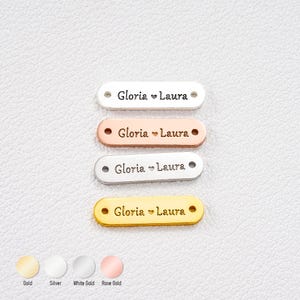 Op de afbeelding: Vier metalen naamlabels in goud, zilver, witgoud en roségoud. Elk label heeft de namen "Gloria" en "Laura" gescheiden door een hartsymbool. De labels zijn ovaal van vorm met een gat aan elk uiteinde.