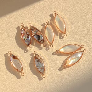 Op de afbeelding: Assortiment van goudkleurige metalen en iriserende schelp hangers. De hangers zijn ovaalvormig en hebben aan elk uiteinde een gat om aan sieraden te bevestigen.
