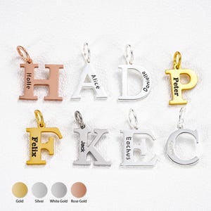 Könnte beinhalten: Eine Auswahl an personalisierten Buchstabenanhängern in Gold, Silber, Weißgold und Roségold. Jeder Anhänger zeigt einen anderen Buchstaben des Alphabets mit eingraviertem Namen. Die Anhänger werden auf weißem Untergrund präsentiert.