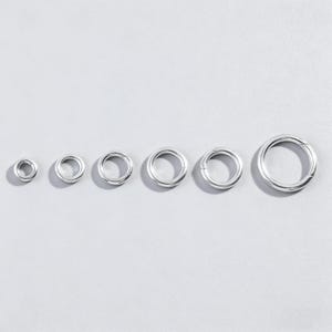 Może przedstawiać: Sześć srebrnych kółek sprężynowych o różnych rozmiarach ułożonych w rzędzie na jasnoszarym tle. Pierścienie, prawdopodobnie metalowe, mają gładką, odblaskową powierzchnię. Rozmiary wahają się od małych do dużych, do biżuterii lub rękodzieła.