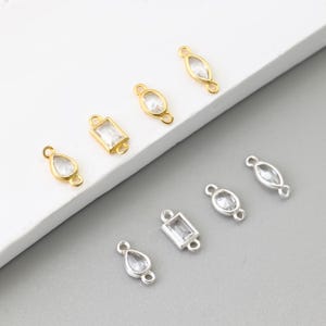 May include: An assortment of jewelry findings in gold and silver tones. The findings feature clear, faceted gemstones in various shapes, including teardrop, oval, and rectangular. Each piece has small loops for attaching to other components.